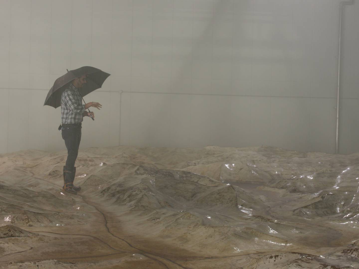 画像: 室内で地形模型の上に立ち、傘を差している男性。ジオラマのような山岳地帯を再現したセットが広がっており、煙や霧が場面