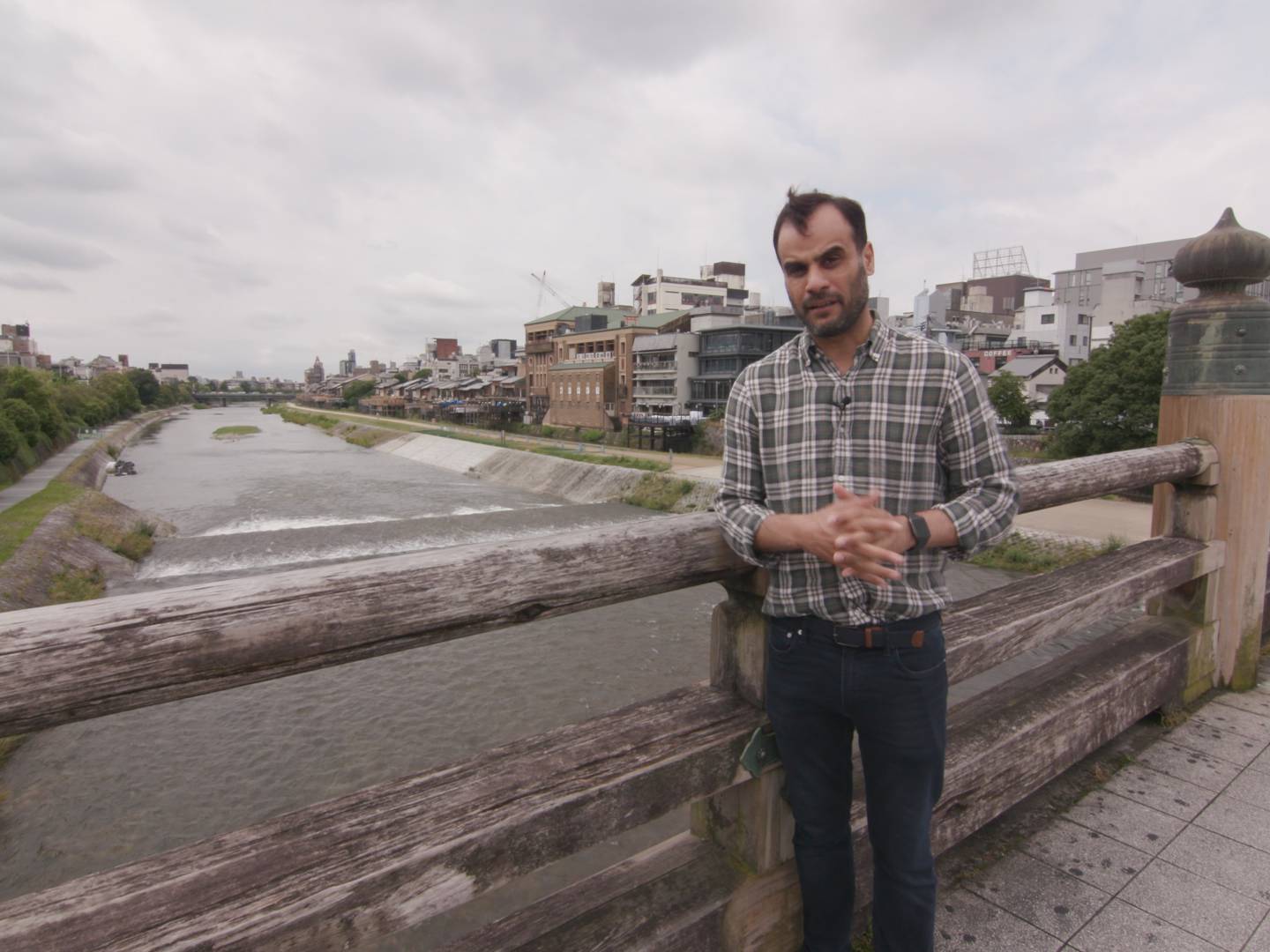 画像: 木製の橋に立つ男性が写っている京都の風景。背景には鴨川とその川岸の建物が見え、曇り空が広がっている。観光スポット、京都