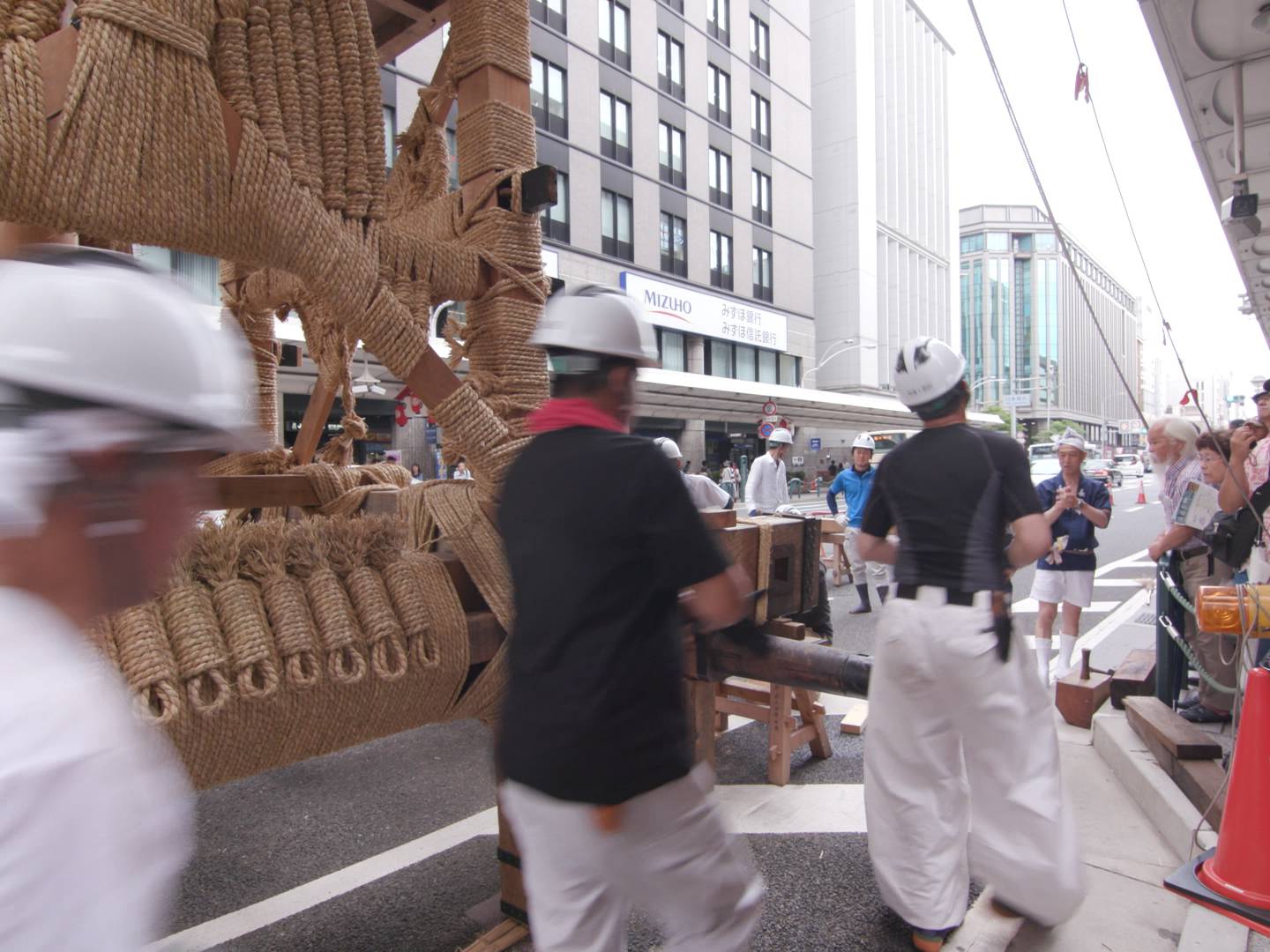 画像: 京都の祇園祭で山鉾建てが行われている様子です。作業員が伝統的な山鉾の設置作業を進めており、周囲には見物客