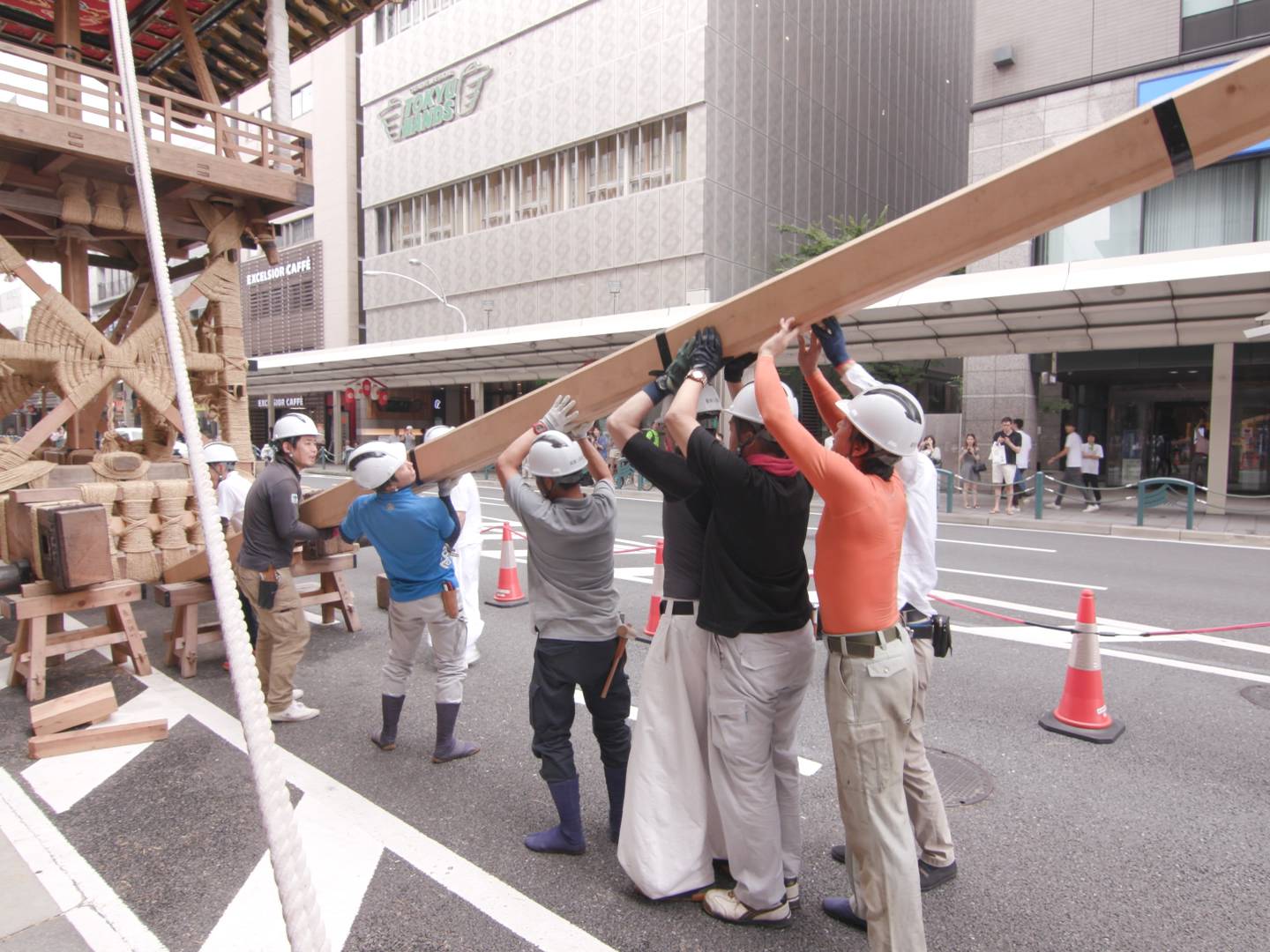 画像: 通りでの祭りの準備中の様子で、作業員が山車を組み立てています。ヘルメットを着用した男性たちが長い木材を持ち上げる