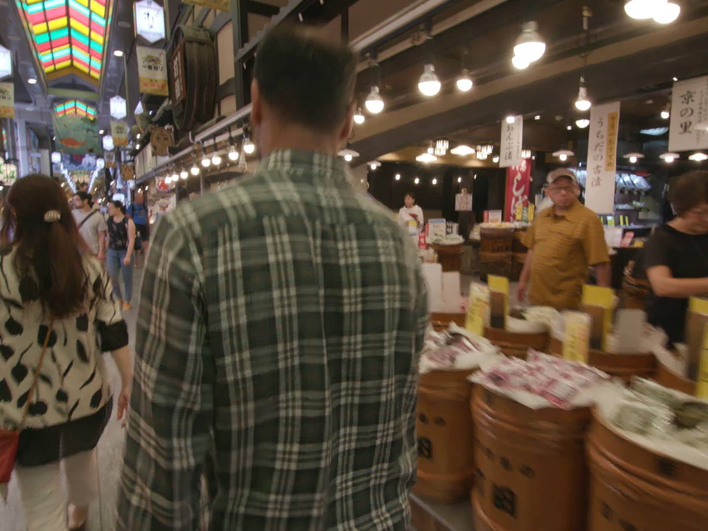 画像: 錦市場の賑わいの場面を撮影した写真です。色とりどりの屋根の下、多くの人が活気ある商店街を歩いています。新鮮な食材