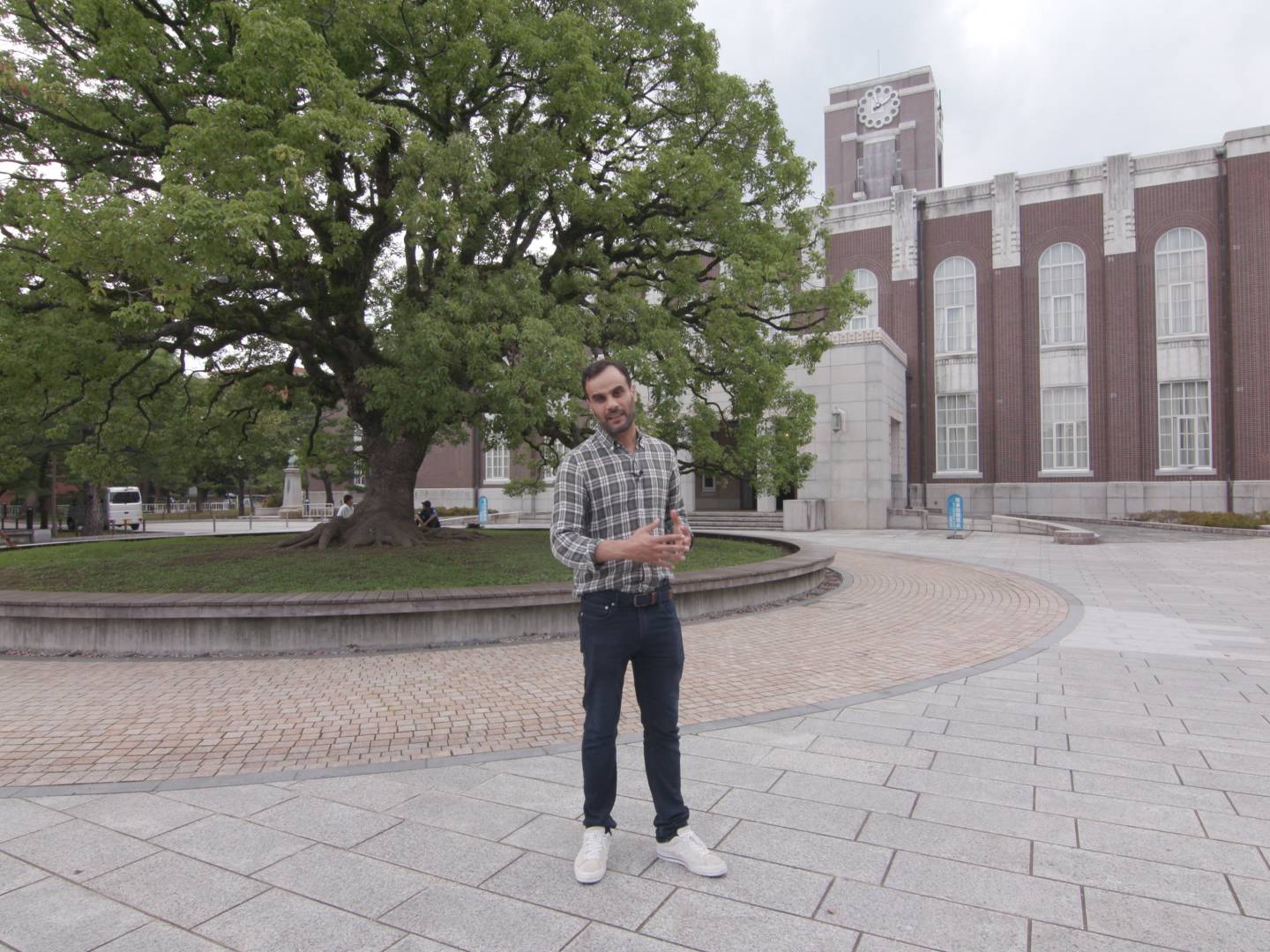 画像: 申し訳ありませんが、人物を識別することはできません。この画像には、大きな木がある広場と古い建物が写っています。場所は大学キャンパスのように見えます。「大学キャンパ