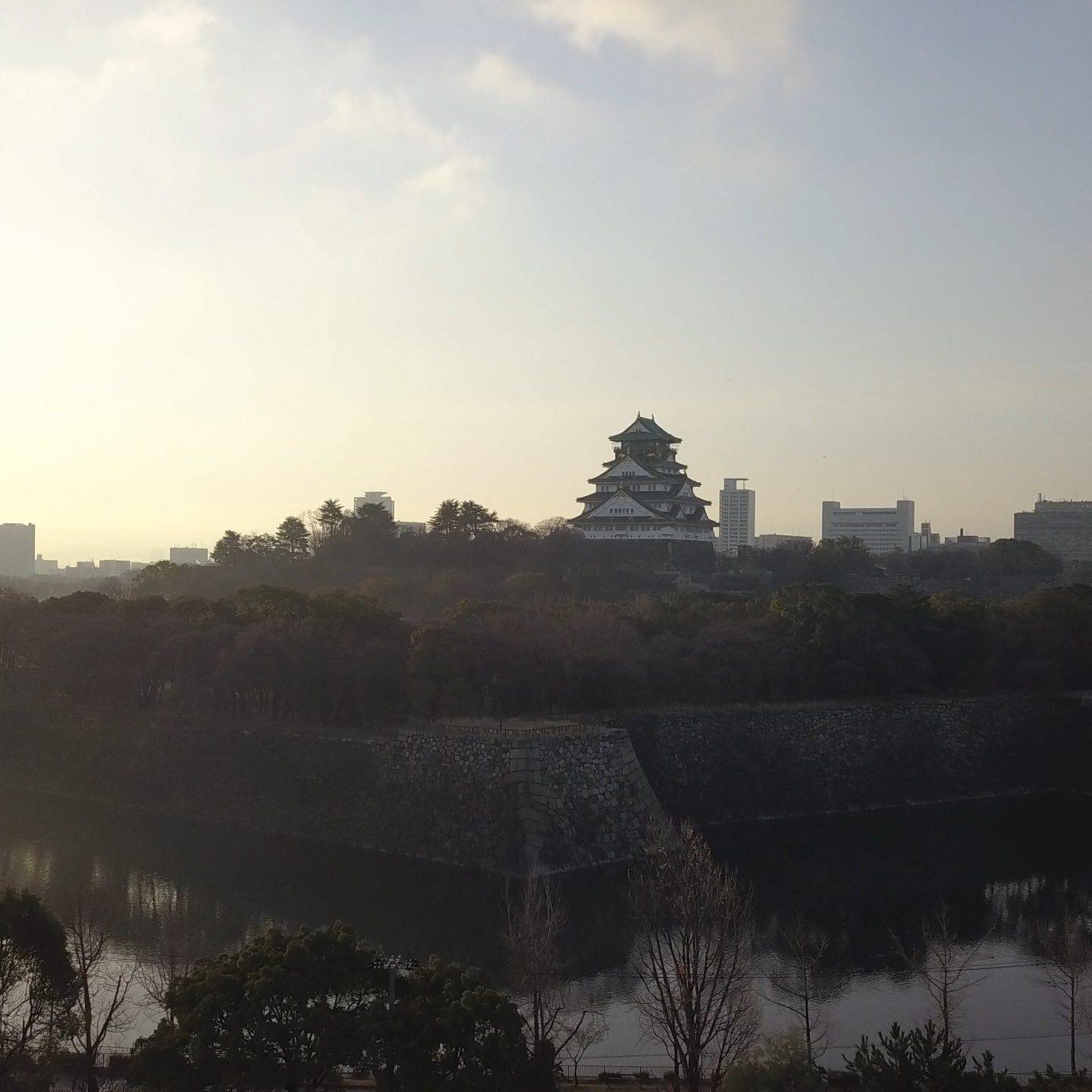 画像: 大阪城が朝日の中に立つ美しい風景。城と周囲の公園が川に映り込み、やわらかな朝の光が広がる。観光、大阪の名所、歴史