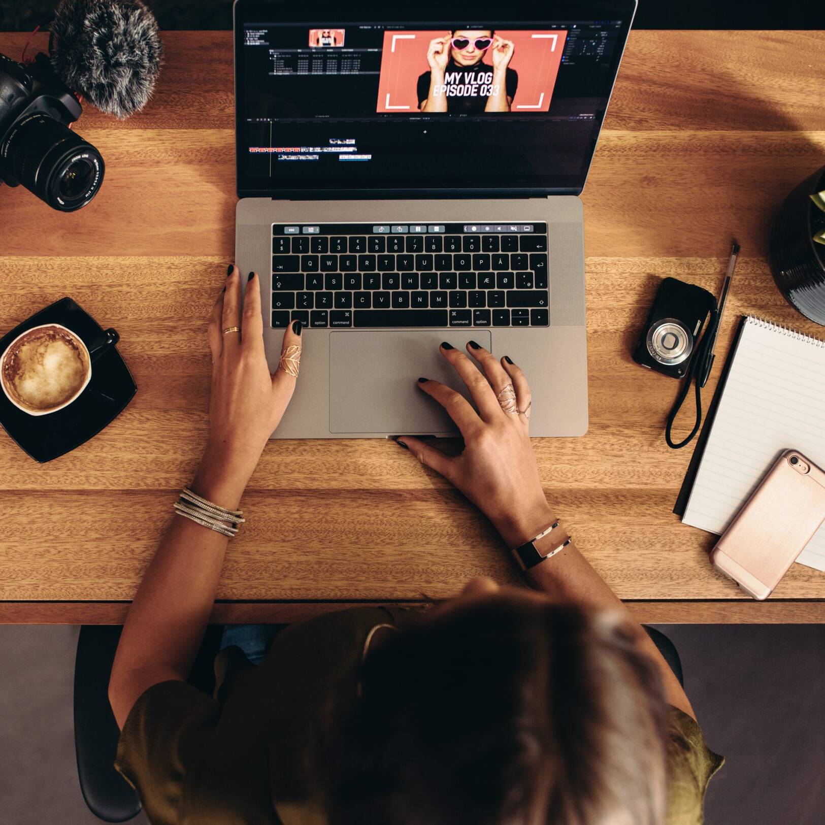 画像: 女性がノートパソコンで動画編集を行うデスクトップシーン。デスクにはカメラ、コーヒー、観葉植物、スマートフォンが配されている。キーワード: 動画編集