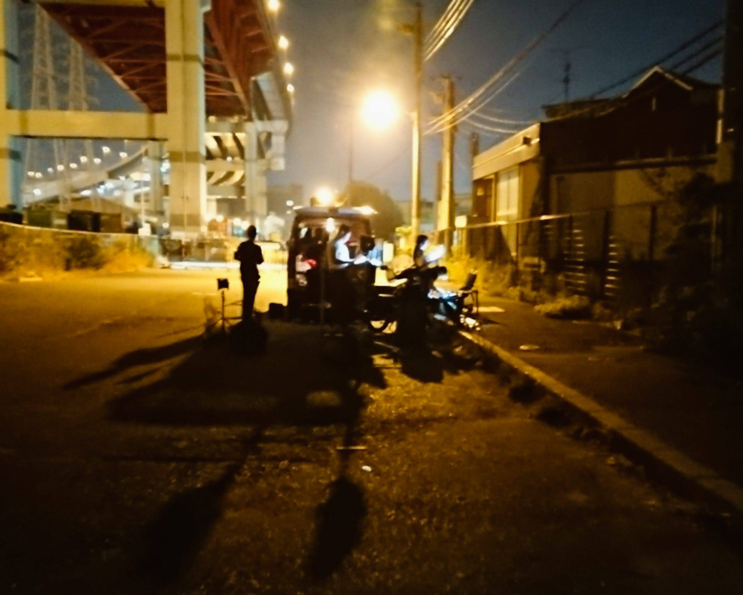 画像: 高速道路の高架下で夜にキャンプをする人々の様子。夜空の下で街灯が照らす中、簡易的なキャンプ椅子とテーブルが見えます。都会