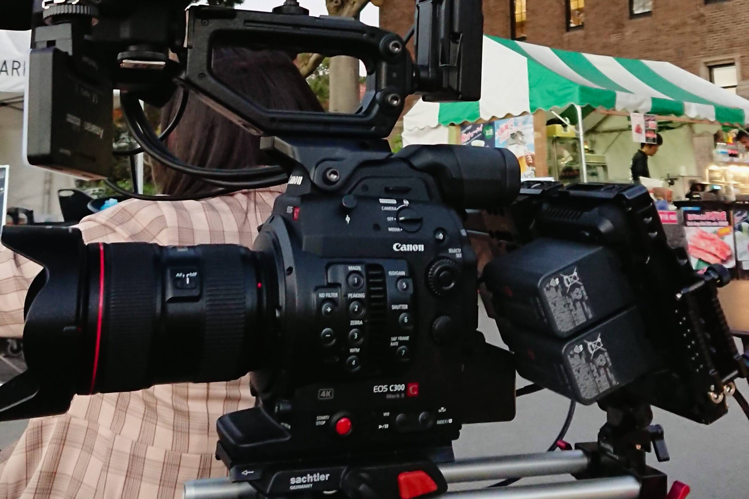 画像: プロの撮影現場でのCanon EOS C300カメラのクローズアップ。背景には屋外イベントのテントが見える。映画製作、ビデオ撮影、プロフェッショナルカメ