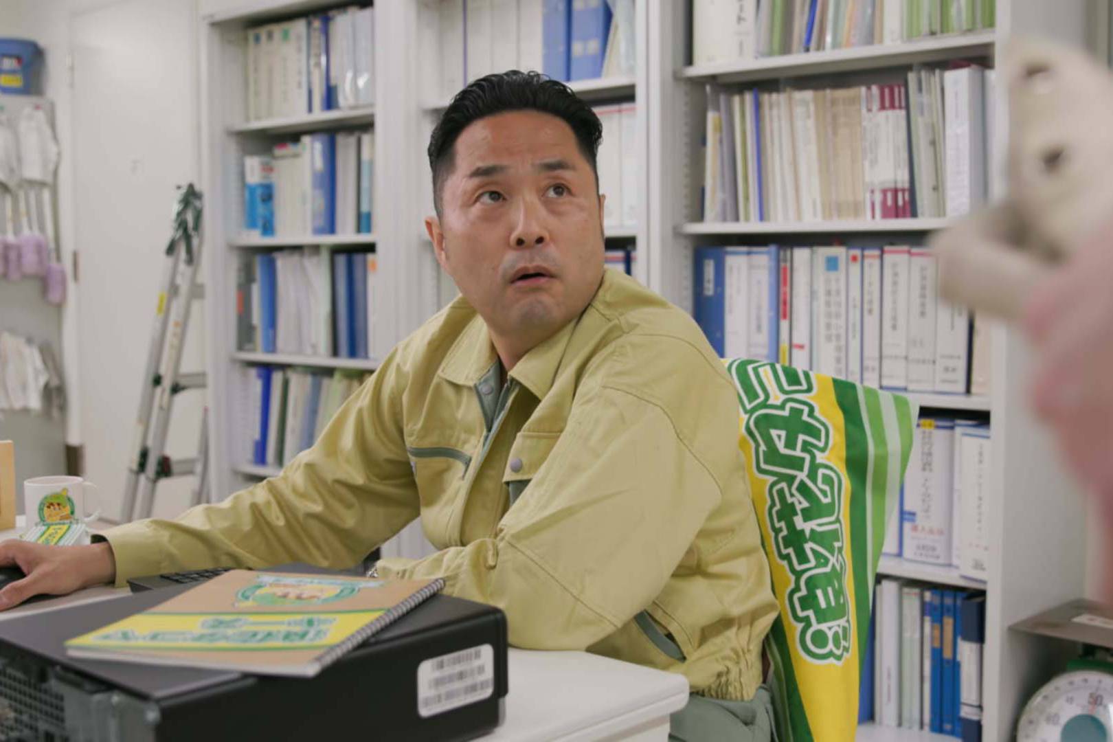 画像: 申し訳ありませんが、この画像に写っている人物についてはわかりません。 オフィス内で作業している男の人がデスクに座っているシーンです。背景には本棚があり、書