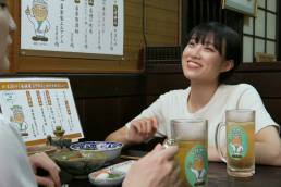 画像: 申し訳ありませんが、人を特定することはできません。この画像は、居酒屋で楽しそうに会話をしている女性が写っているシーンです。テーブルにはビールジョッキや料理が並