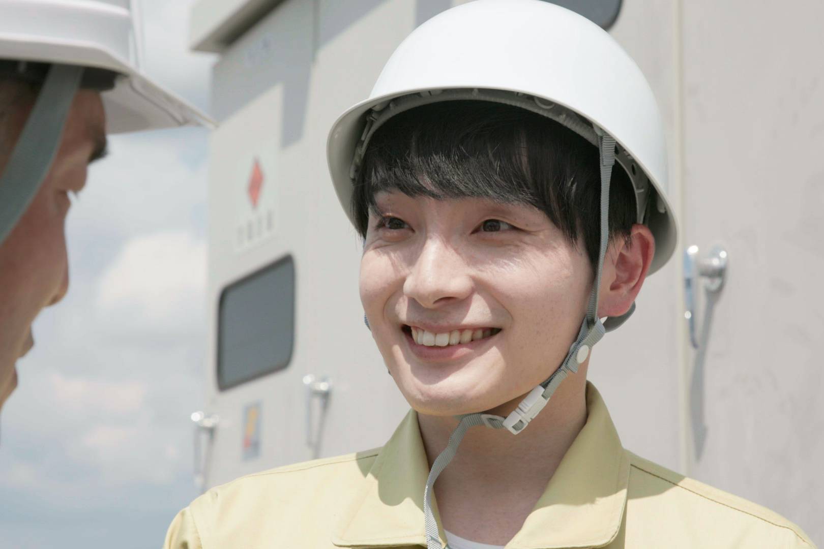画像: 申し訳ありませんが、この画像の特定の人物を認識することはできません。ただし、工事現場での安全ヘルメットを着用した作業員が笑顔で会話しているシーン