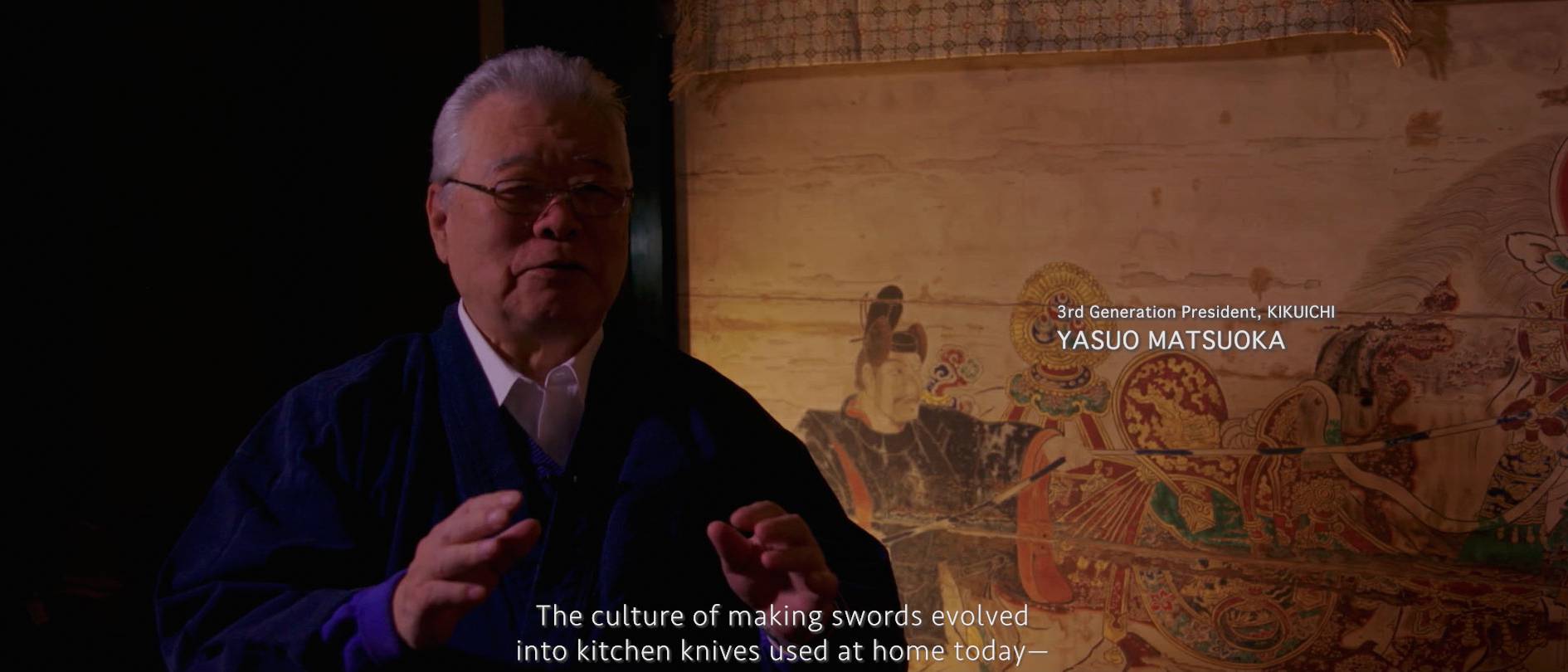 画像: 申し訳ありませんが、画像内の人物を認識することはできません。この画像は、日本の刀鍛冶の伝統について話す人物が描かれており、背景には日本絵画が見ら