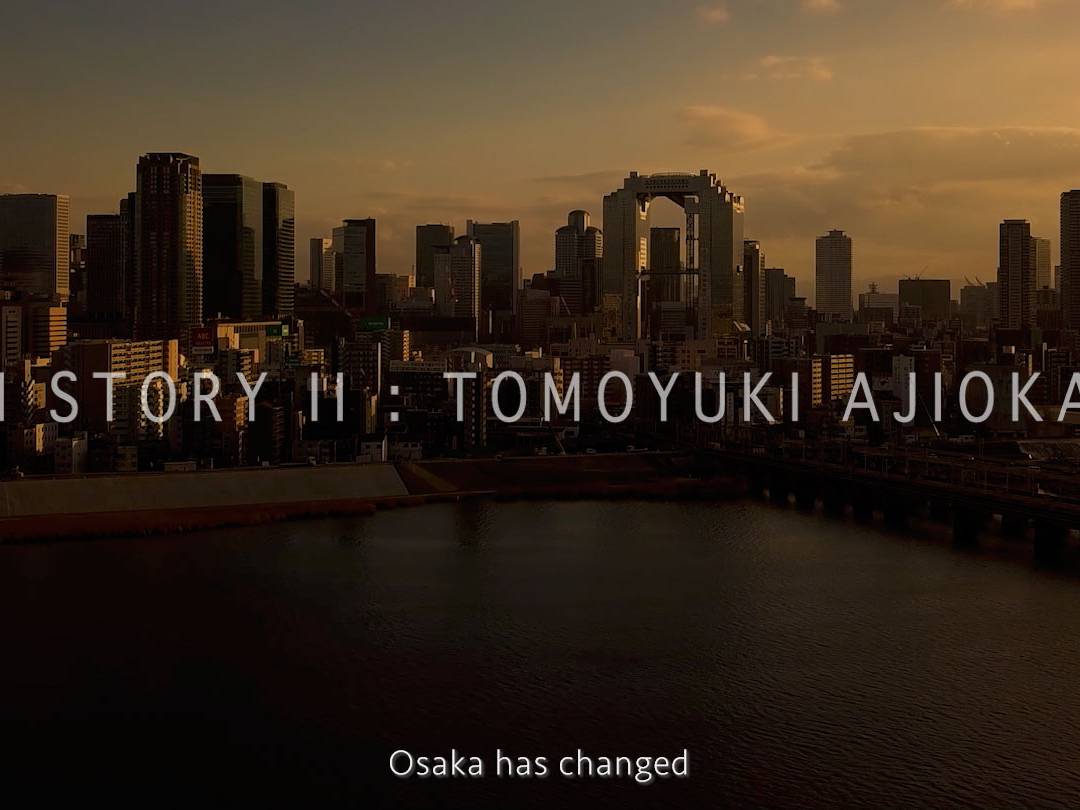 画像: 大阪の夕暮れの都市景観を背景にした「KIKUICHI STORY II: TOMOYUKI AJIOKA, GRIND」というテキストが表示された画像です。淀川の水面と都市の