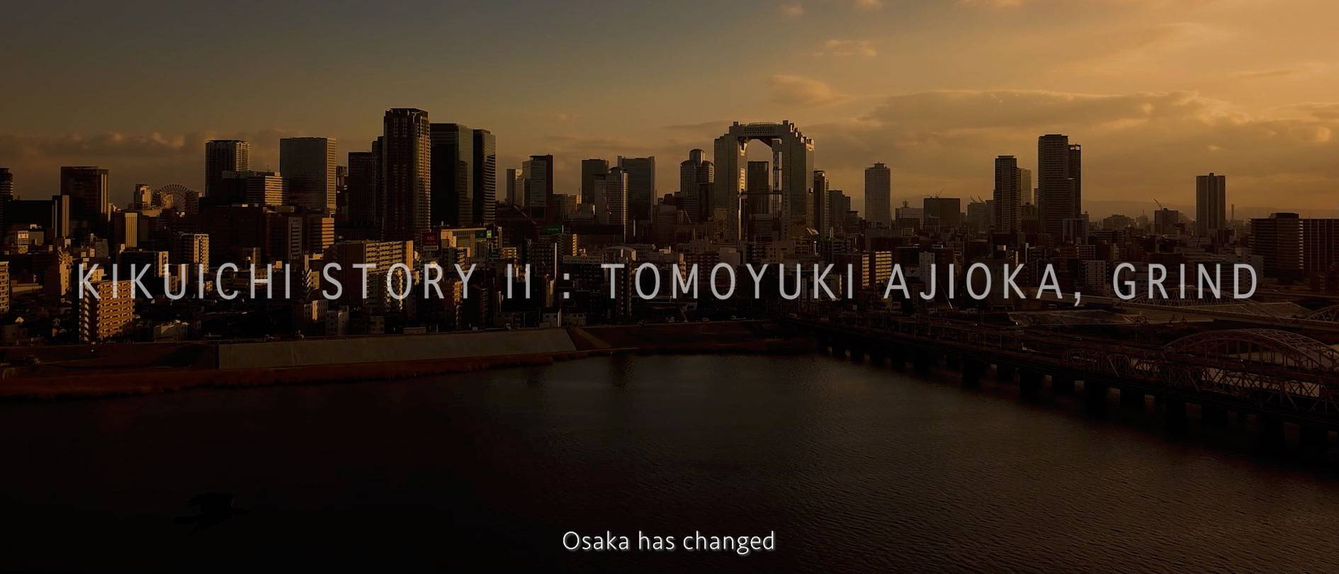 画像: 大阪の夕暮れの都市景観を背景にした「KIKUICHI STORY II: TOMOYUKI AJIOKA, GRIND」というテキストが表示された画像です。淀川の水面と都市の