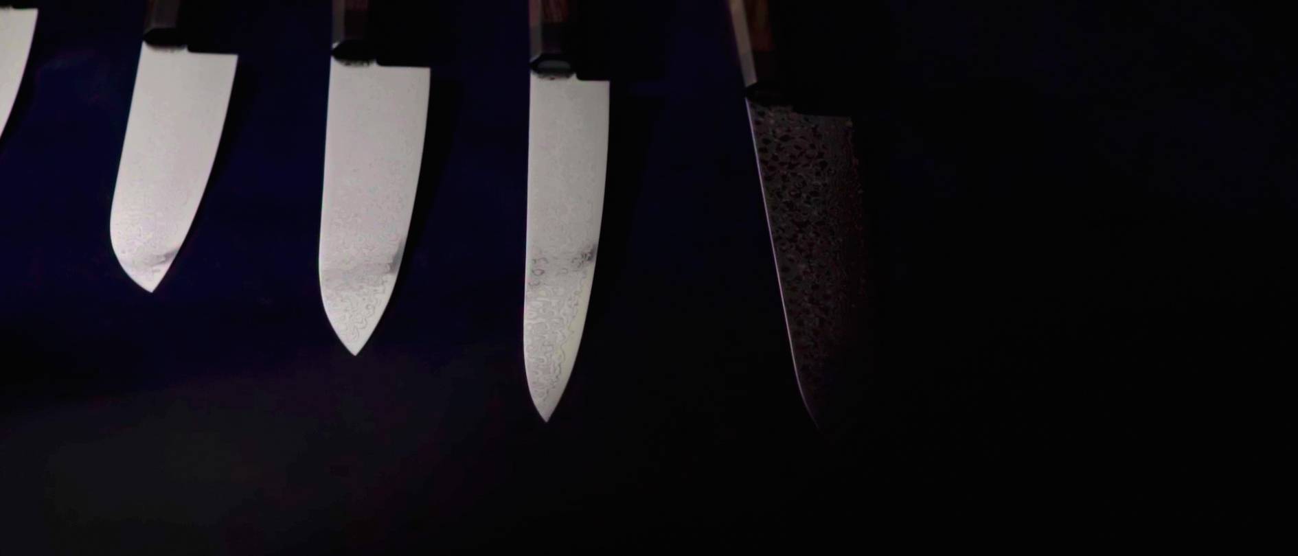 画像: この画像は、暗い背景に並んだ4本のナイフの刃部分をクローズアップで捉えています。ナイフは異なる質感や模様を持ち、それぞれの