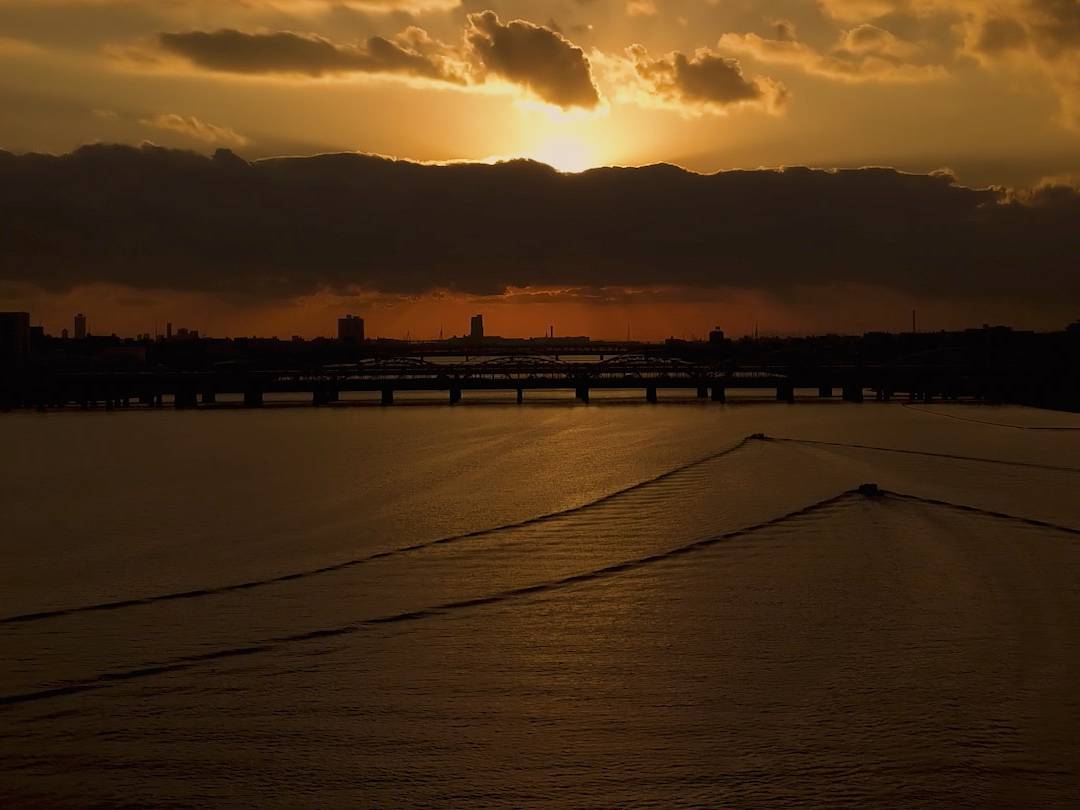 画像: 黄昏時の川の景色です。沈む夕日が雲の間から顔を出し、穏やかな水面に光を投げかけています。橋とシルエットが広が