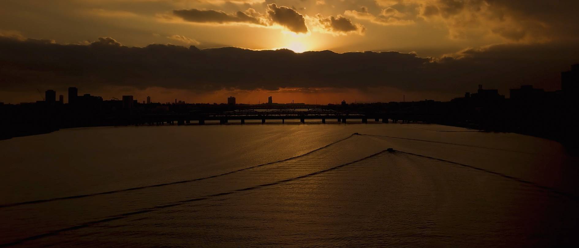 画像: 黄昏時の川の景色です。沈む夕日が雲の間から顔を出し、穏やかな水面に光を投げかけています。橋とシルエットが広が