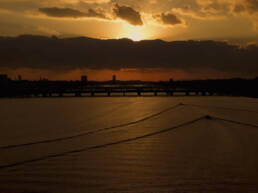 画像: 黄昏時の川の景色です。沈む夕日が雲の間から顔を出し、穏やかな水面に光を投げかけています。橋とシルエットが広が